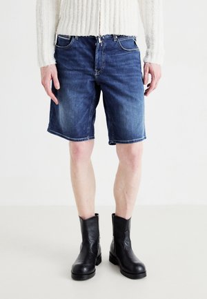 Mand iført midtvaske blå denimshorts, hvid tekstureret sweater og sorte læderstøvler til midt på læggen, stående mod en ensfarvet hvid baggrund.