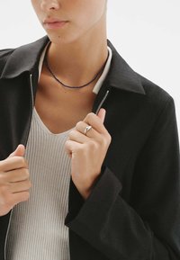 Chaqueta negra con cremallera y cuello, combinada con una blusa ligera de canalé. Incluye un collar de cuentas azules y un anillo de plata.
