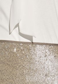 Weißes Oberteil mit Schleifendetail, über einem goldenen Paillettenbereich. Die Pailletten erzeugen eine strukturierte, glänzende Oberfläche im Kontrast zu dem glatten Stoff.