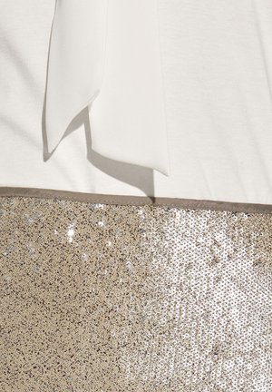 Weißes Oberteil mit Schleifendetail, über einem goldenen Paillettenbereich. Die Pailletten erzeugen eine strukturierte, glänzende Oberfläche im Kontrast zu dem glatten Stoff.