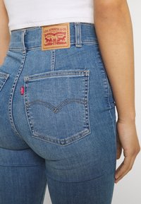 Jeans de mezclilla azules de cintura alta que presentan un parche de cuero con el logo, bolsillos traseros con un diseño de costura curvada y una etiqueta roja en el costado.