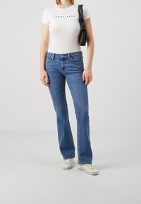 Camiseta blanca de manga corta con el logo "TOMMY JEANS", combinada con vaqueros acampanados de color azul claro. Bolso negro al hombro y zapatillas blancas.