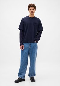 Granatowy t-shirt z krótkim rękawem i kieszonką na piersi, noszony na ciemnym, długim rękawem, zestawiony z luźnymi, jasnoniebieskimi dżinsami i czarnymi butami.