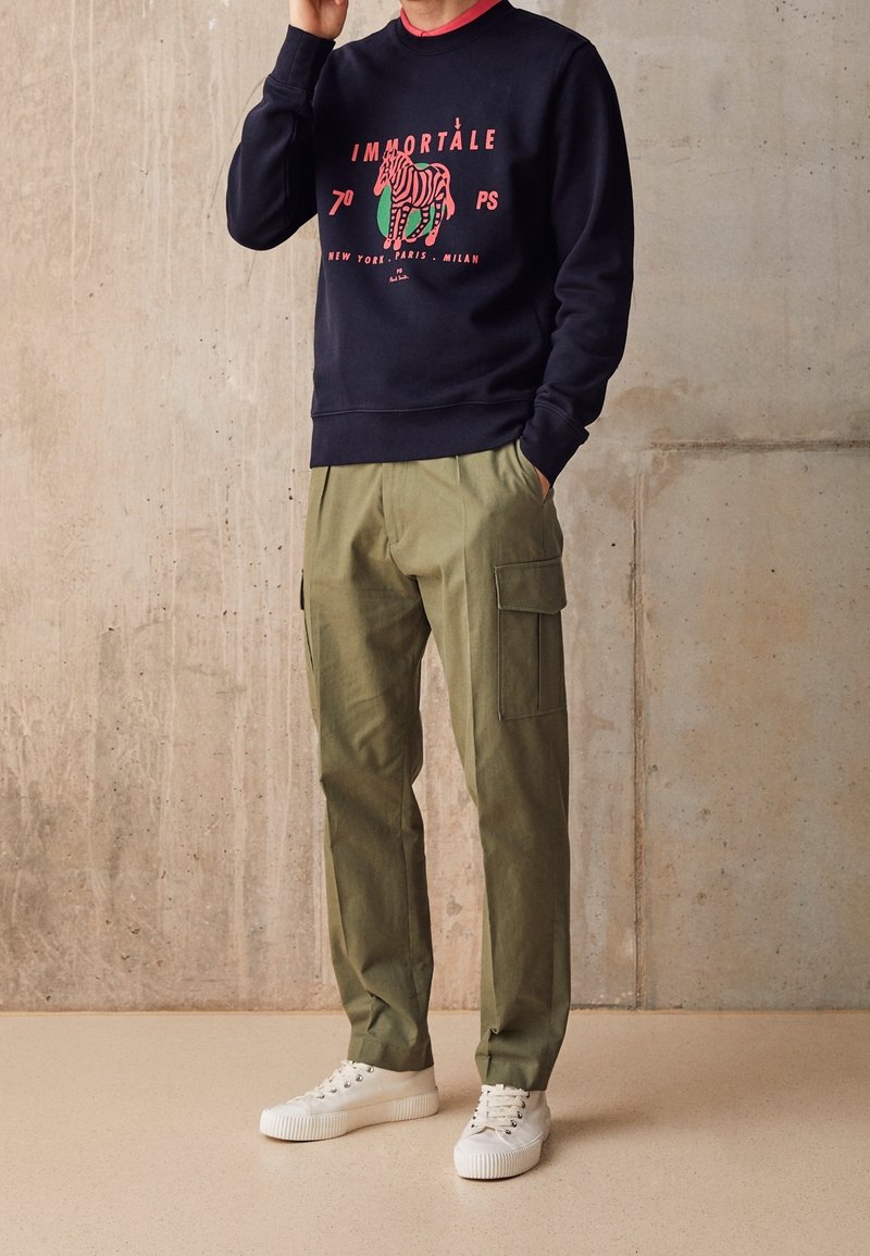 Personne portant un sweat-shirt noir avec un graphisme zèbre rouge et du texte, un pantalon cargo vert olive et des baskets blanches, debout contre un mur en béton.