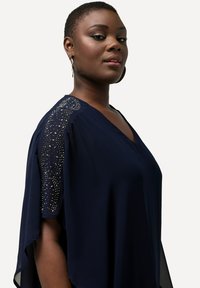 Blusa blu navy con scollatura a V e maniche corte, dotata di dettagli decorativi sulle spalle, realizzata in un tessuto leggero.