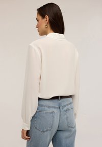 Blouse blanche à manches longues, avec un col et des poignets boutonnés, associée à un jean taille haute bleu clair, agrémenté d'une ceinture marron et de poches.