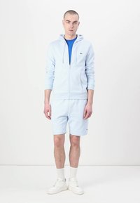 Ανοιχτό μπλε φούτερ με φερμουάρ και κορδόνια, συνδυασμένο με κοντό παντελόνι ιδίου χρώματος. Μπλε μπλούζα από κάτω. Λευκά αθλητικά παπούτσια και λευκές ρίγες κάλτσες.