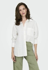 White linen shirt with a button-down front, long sleeves, and a single chest pocket. Paired with light green wide-leg pants.