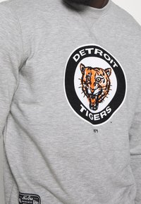 Šedá mikina s kulatým černobílým logem zobrazujícím hlavu tygra a textem "DETROIT TIGERS". Měkká tkanina, kulatý výstřih a postranní štítek.