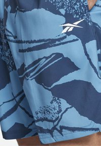 Niebieskie spodenki sportowe z ciemnymi abstrakcyjnymi wzorami i białym logo Reebok w górnym prawym rogu, noszone przez osobę.