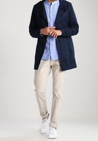 Cappotto impermeabile navy con chiusura a bottoni, camicia azzurra chiaro con bottoni, chinos beige e sneakers bianche. Tessuto liscio e vestibilità su misura.