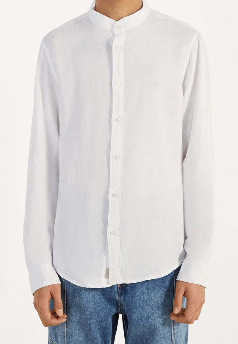 Uomo che indossa una camicia bianca semplice a maniche lunghe con colletto alla cinese e jeans in denim blu, in piedi con le braccia rilassate.