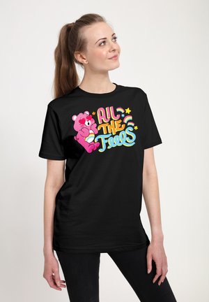 Jonge vrouw draagt een zwart shirt met een kleurrijke cartoonbeer en de tekst "All the Feels" met sterren en regenbogen, kijkt omhoog en glimlacht.