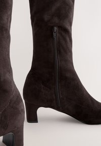 Stivali al ginocchio in camoscio marrone scuro con cerniera laterale, caratterizzati da una finitura testurizzata e un tacco modesto. Bordi lisci e forma su misura.