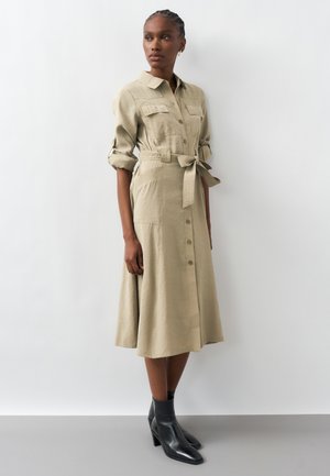 Γυναίκα που στέκεται, φορώντας ένα μπεζ μεσαίο φόρεμα με κουμπιά, τυλιγμένα μανίκια και δεμένη ζώνη στη μέση, σε συνδυασμό με μαύρα μποτάκια αστραγάλου.