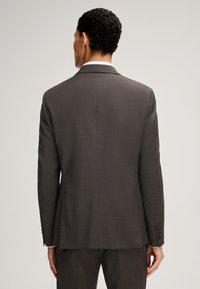 Uomo visto da dietro che indossa un giacca scura grigia e pantaloni coordinati su uno sfondo chiaro e semplice.