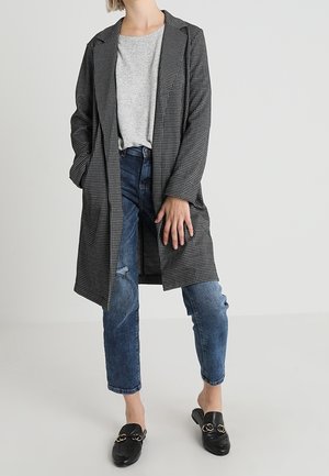 Vrouw met een grijze geruite lange jas, lichtgrijs T-shirt, blauwe gescheurde spijkerbroek en zwarte instaploafers, staand tegen een effen achtergrond.