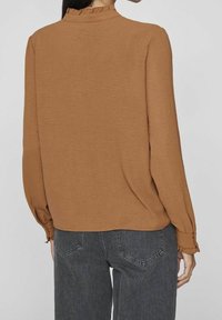 Brun långärmad topp med strukturerad finish och rynkad halsringning. Kombinerad med högmidjade, mörkgrå jeans.