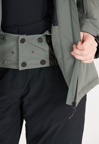 Grüne Außenjacke mit einer schwarzen Innenschicht, ausgestattet mit einem Druckknopfverschluss, elastischem Bund und Reißverschlusstaschen an den Seiten, kombiniert mit dunklen Hosen.