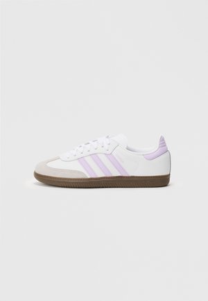 Sneakers Adidas Samba blancs et beiges avec des rayures violettes pastel et une semelle en caoutchouc, présentés de profil sur un fond gris clair.