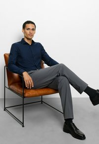 Dunkelblaues Hemd mit Knöpfen, graue maßgeschneiderte Hose, schwarze Schuhe, sitzt auf einem braunen Ledersessel mit Metallrahmen, minimalistisch gestalteter Hintergrund.