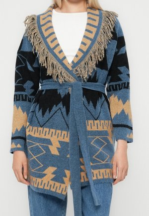 Cardigan en tricot avec motifs géométriques bleus, noirs et beiges, col châle à franges, ceinture assortie nouée à la taille, porté sur un top blanc.