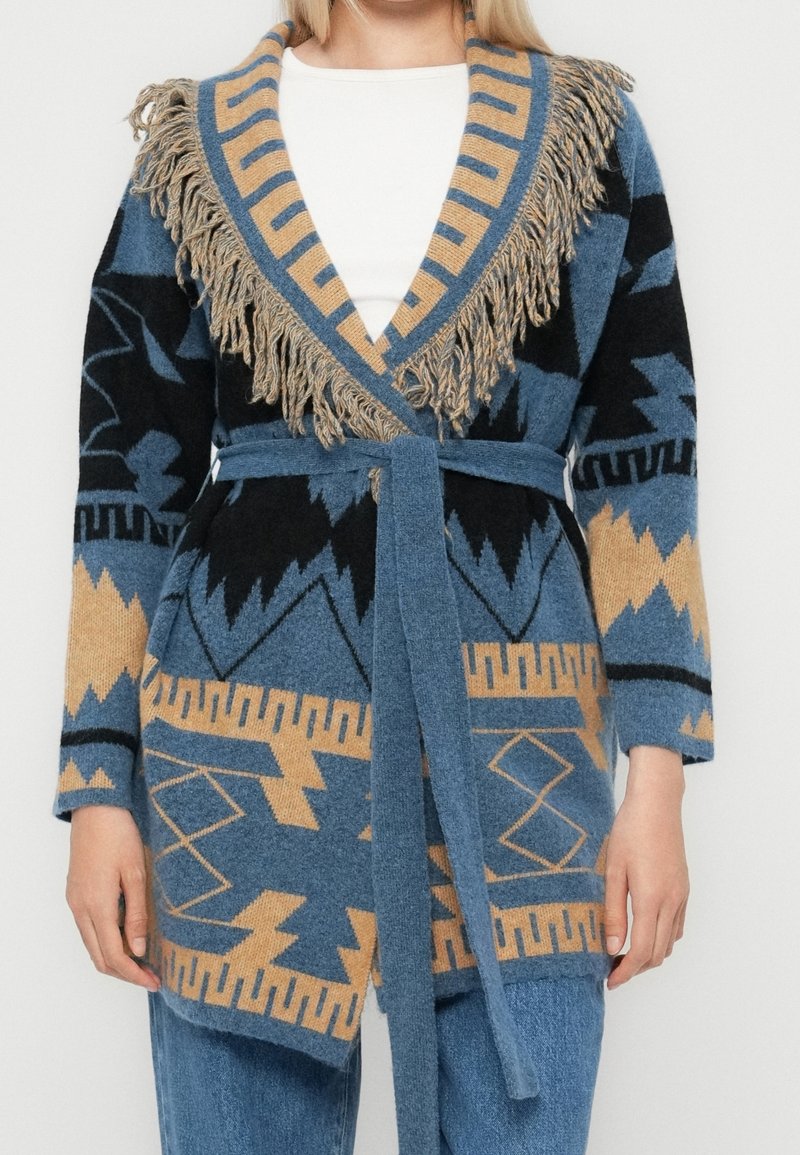 Cardigan en tricot avec motifs géométriques bleus, noirs et beiges, col châle à franges, ceinture assortie nouée à la taille, porté sur un top blanc.