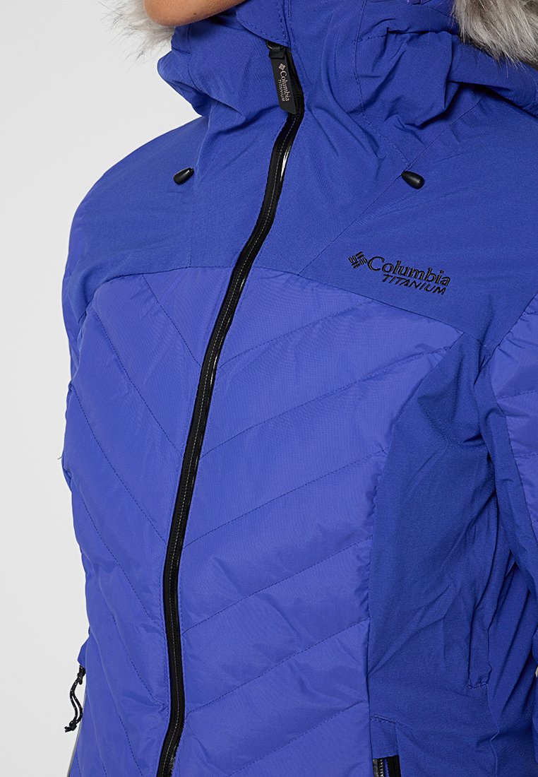 Veste matelassée violette avec un col montant, fermeture éclair à l'avant, motif chevron texturé et logo Columbia Titanium sur la poitrine.