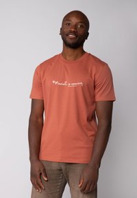 T-shirt en coton couleur rouille avec des manches courtes et un col ras du cou, comportant un texte imprimé blanc qui dit "Mariah arrive."