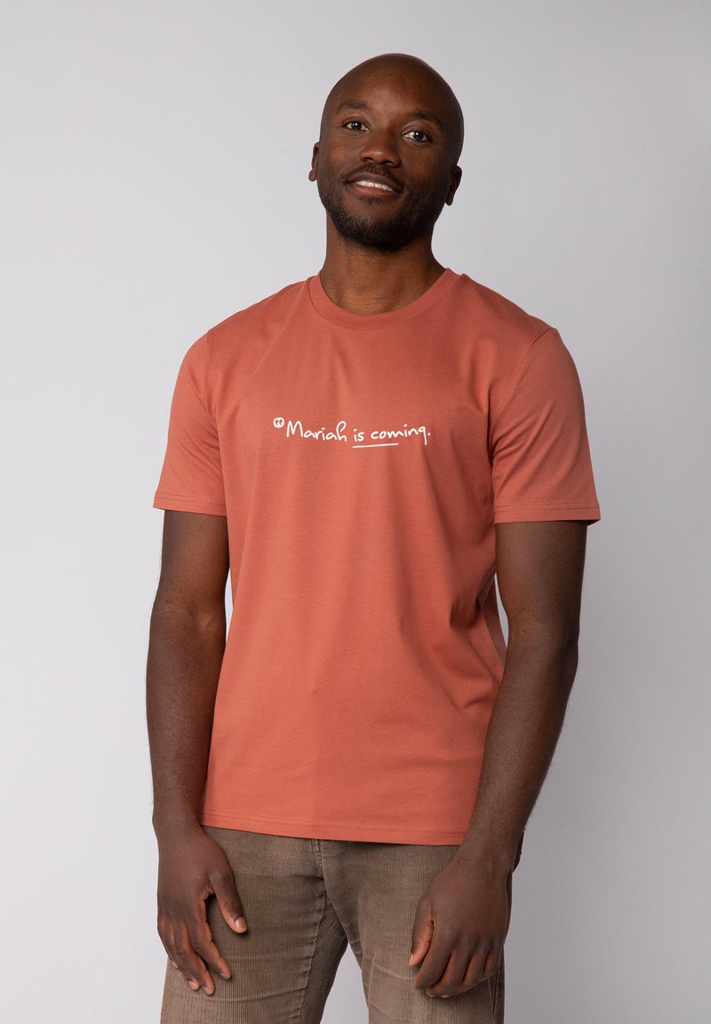 T-shirt en coton couleur rouille avec des manches courtes et un col ras du cou, comportant un texte imprimé blanc qui dit "Mariah arrive."