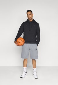 Svart hettegenser med et okse-motiv og frontlomme, kombinert med grå shorts. Modellen holder en oransje basketball og har på seg hvite sko med svarte detaljer.