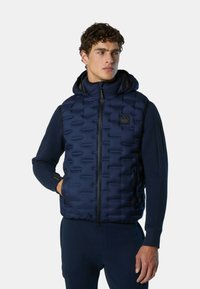 Marineblauer Puffer-Weste mit Kapuze, gestepptem Material, Reißverschluss vorn und seitlichen Taschen. Über einem passenden marineblauen Oberteil getragen.