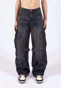 Άτομο φορώντας μαύρο cargo παντελόνι με φαρδιά πόδια και πολλές μεγάλες τσέπες, καθώς στέκεται απέναντι σε πολύχρωμο φόντο.