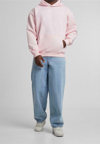 Person som bär en ljusrosa huvtröja, lösa ljusblå jeans och vita sneakers, står med ena handen i huvtröjans ficka.