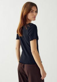 Melna ribota īsām piedurknēm blūze ar viļņotu apakšmalu un pieguļošu dizainu. Valkāta ar brūnām biksēm, kas parāda gludu tekstūru un pieguļošu siluetu.