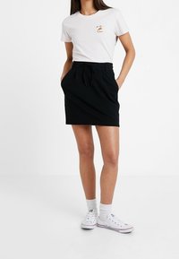 Gonna nera con tasche frontali e posteriori, realizzata in tessuto liscio, abbinata a una camicia bianca con stampa grafica, indossata con sneaker bianche.