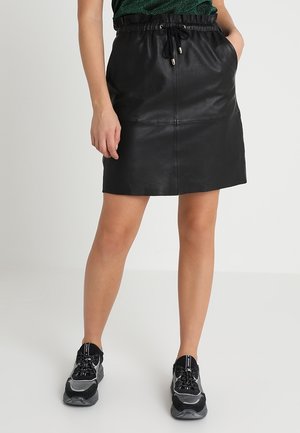 Femme portant une mini-jupe en cuir noir avec une ceinture élastique et des baskets noires et grises à lacets, debout sur un sol blanc.