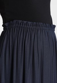 Sukně v námořnické modři s elastickým pasem má splývavý, plisovaný design. Tkanina vypadá hladce s mírným leskem.