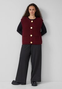 Burgunderfarbener Strickpullover mit goldenen Knöpfen, zwei Vorderaschen und einem runden Halsausschnitt, kombiniert mit schwarzen weit geschnittenen Hosen mit Nadelstreifen.