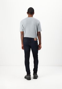 Άνδρας γυρισμένος με την πλάτη, �φορώντας γκρι μπλουζάκι, σκούρα skinny jeans με δερμάτινη ετικέτα και μαύρα αθλητικά παπούτσια, σε ουδέτερο λευκό φόντο.