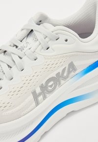 Baskets de sport blanches avec un dessus en mesh, logo HOKA argenté et semelle en dégradé bleu. Présente un design léger et des détails texturés.