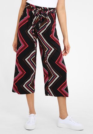 Kobieta nosząca czarne, krótkie spodnie z szerokimi nogawkami, wielokolorowe paski w zygzak oraz przewiązany pasek w talii, w połączeniu z białymi trampkami.