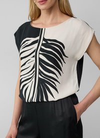 Blouse noir et crème à manches courtes, présentant un design de feuilles audacieux sur le devant, fabriquée en tissu lisse avec une coupe décontractée.
