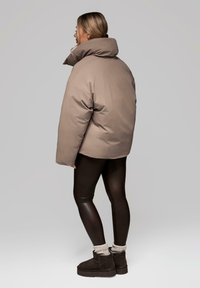 Beżowa kurtka puchowa z wysokim kołnierzem, szerokimi ramionami i gładką fakturą, zestawiona z czarnymi legginsami oraz botkami za kostkę i szarymi skarpetkami.