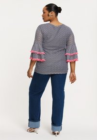 Bluse mit blauem Muster und rosa Pompon-Akzenten, ausgestelltem Ärmel, kombiniert mit dunkelblauen, hochgekrempelten Jeans und weißen Sandalen.