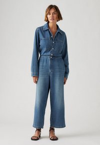 Combinaison en denim bleu clair au design de chemise à col, manches longues, deux poches poitrine et silhouette à jambes larges ; associée à des sandales noires.