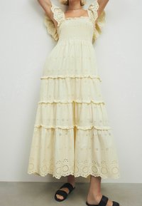 Robe jaune de longueur genou à niveaux avec broderie anglaise, manches volantées et corsage smocké, assortie à des sandales noires à brides.