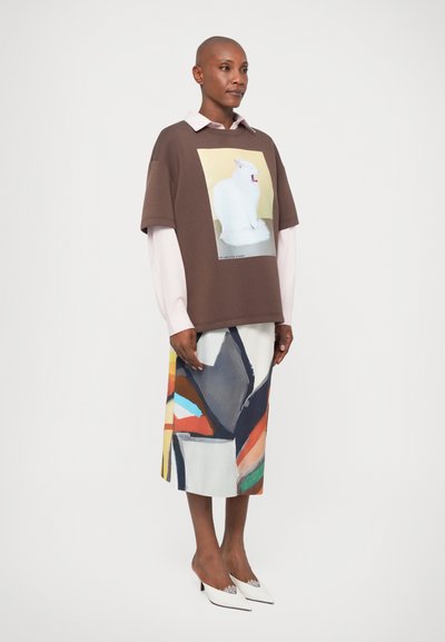 Brązowa, oversize'owa koszulka z grafiką białego kota; nałożona na jasnoróżową koszulę. Kolorowa, wzorzysta spódnica midi i białe obcasy.