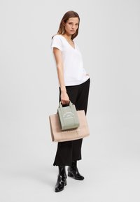 Leinen-Shopper in hellem Beige mit weißen Text und einer grünen Mini-Tasche darauf. Das Model trägt ein weißes V-Ausschnitt-T-Shirt und schwarze Hosen.