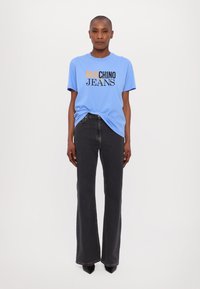 Gaiši zils īsām piedurknēm T-krekls ar melnu un oranžu grafisko tekstu "MOSCHINO JEANS", apvienots ar augsta jostasvietas melnam zvērveida džinsiem.